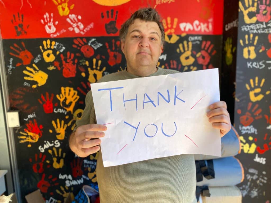 Josh Peachey holds a thank you sign in front of a wall painted with the colours of the Aboriginal flag